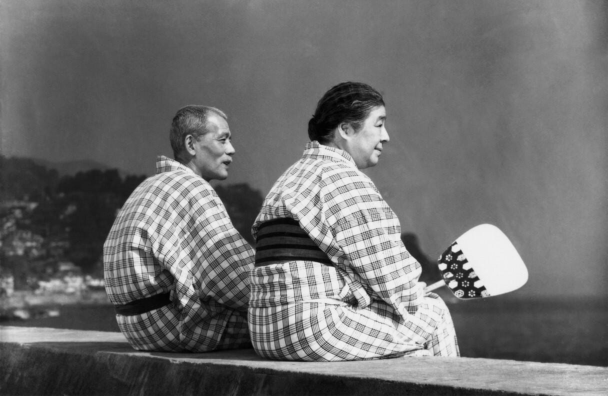 A still from Tokyo Story, with two characters sitting on a ledge, looking out at the ocean.